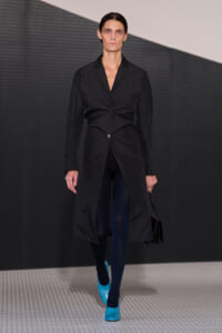 Runway model wearing a long black coat with a twisted front button, navy tights, and bright cyan heels, carrying a black handbag.