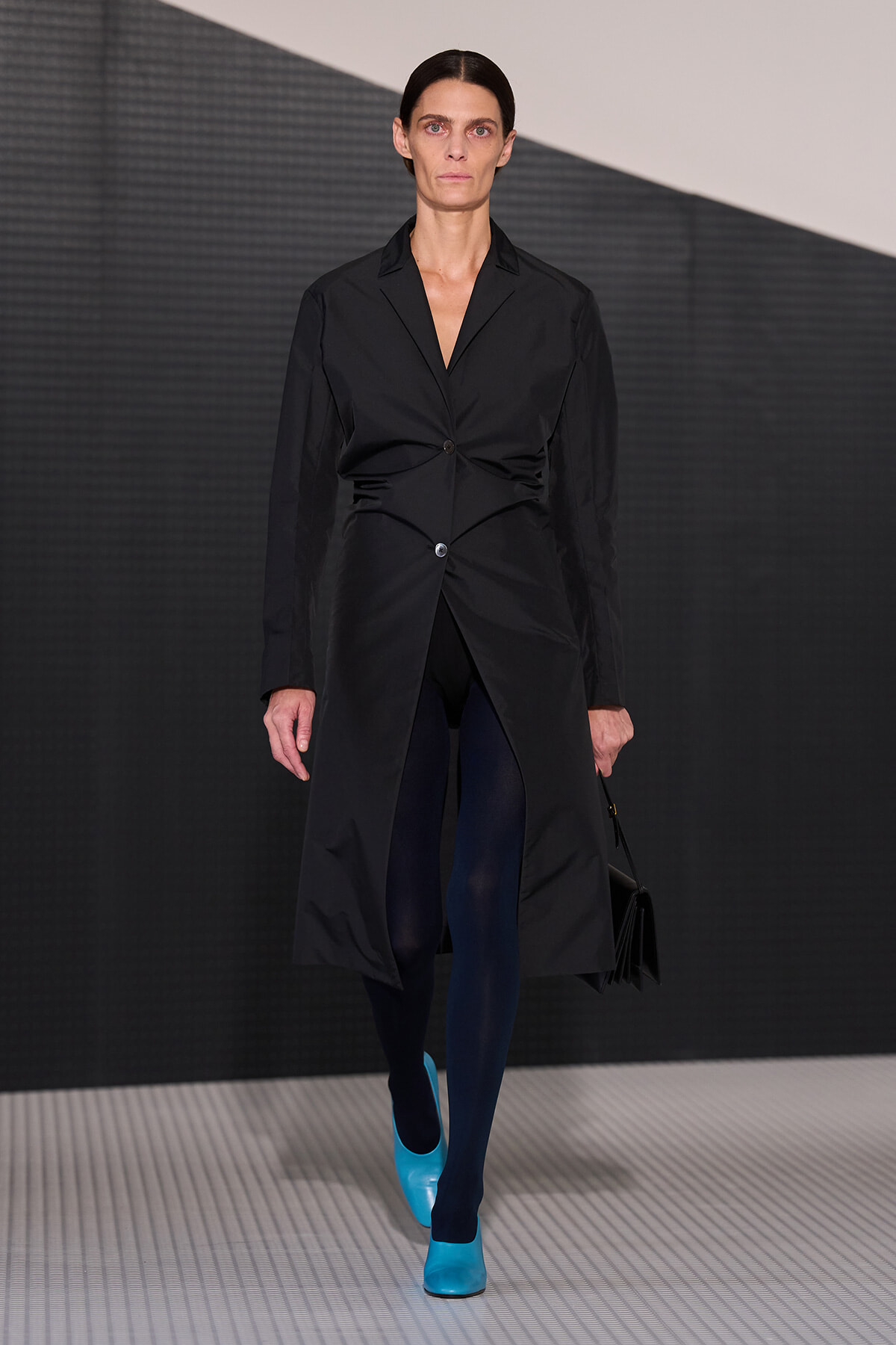 Runway model wearing a long black coat with a twisted front button, navy tights, and bright cyan heels, carrying a black handbag.