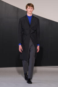 Runway model wearing a knee‑length black coat over a blue knit top and gray trousers, black lace‑up shoes, walking on a patterned catwalk.