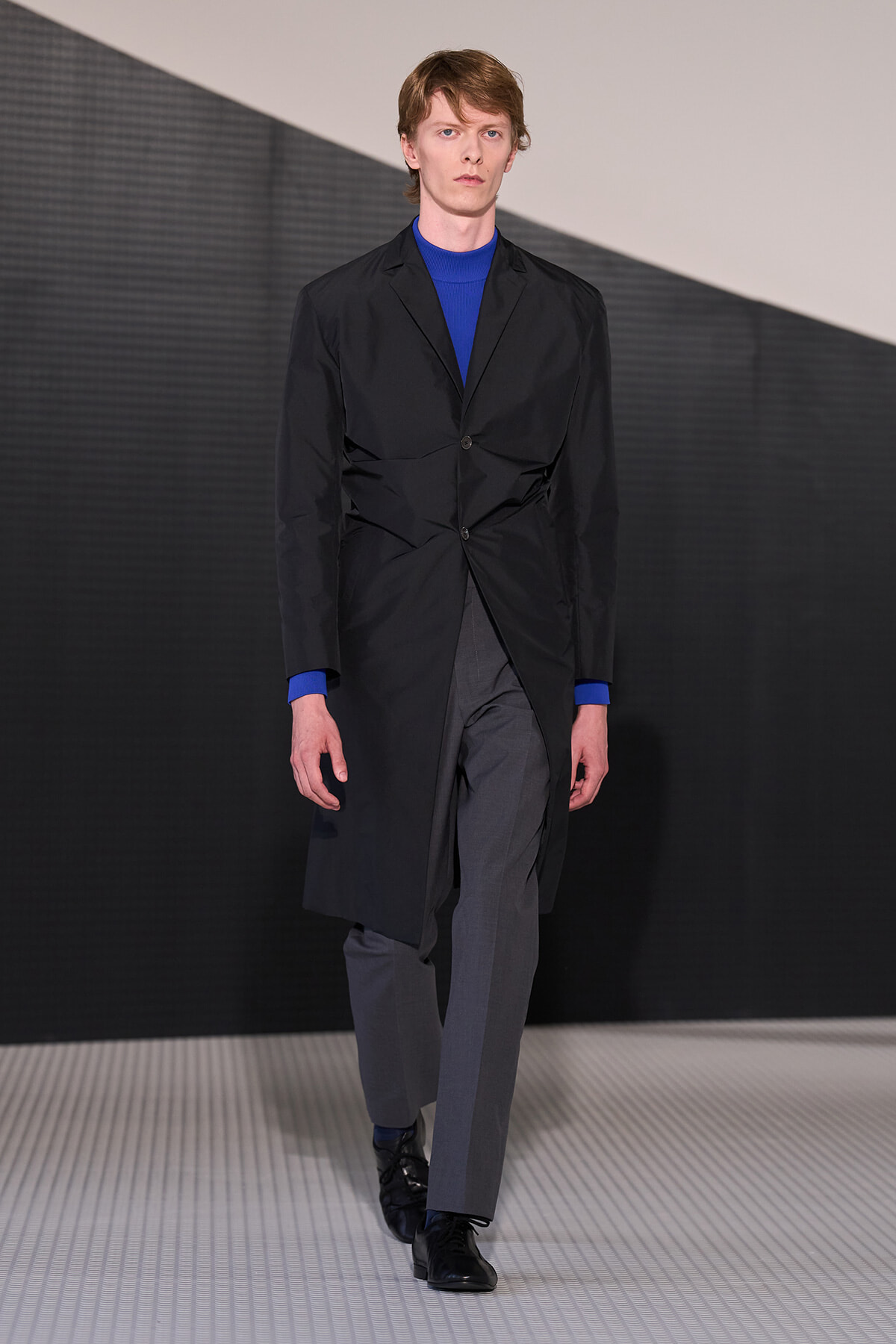 Runway model wearing a knee‑length black coat over a blue knit top and gray trousers, black lace‑up shoes, walking on a patterned catwalk.