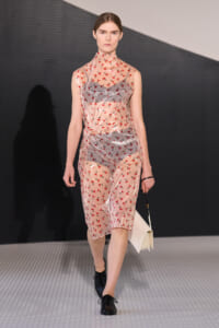 Model walking a runway in a sheer pink floral dress with red blossoms, black shoes, and a pale clutch.