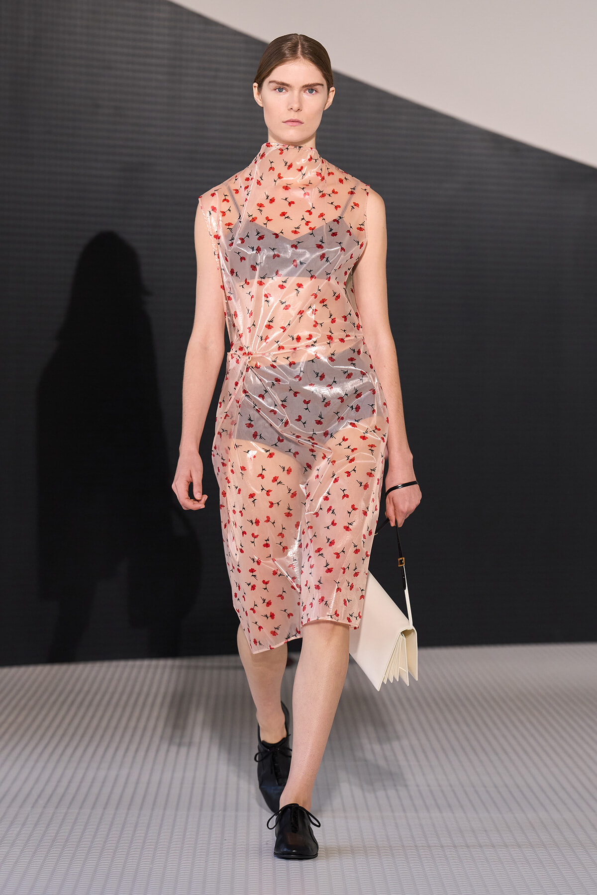 Model walking a runway in a sheer pink floral dress with red blossoms, black shoes, and a pale clutch.