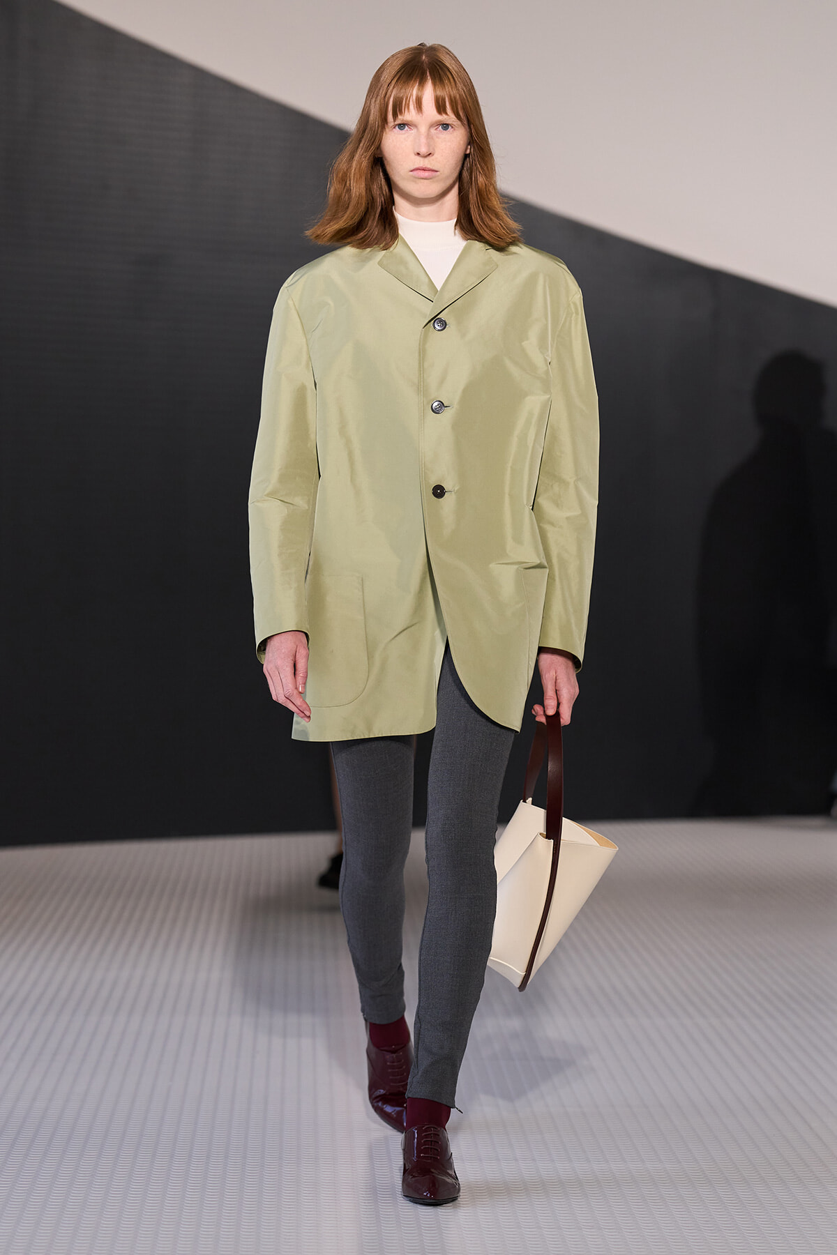 Model walking a runway in a pale green oversized coat over gray leggings, burgundy shoes, and a beige handbag with a dark brown handle.