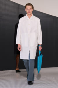 Male model walks a fashion runway in a long white coat, blue-grey pants, and black shoes, carrying a bright blue tote bag.
