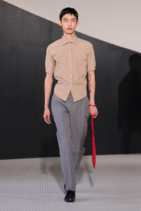 Male model on a runway wearing a beige short-sleeve button-up shirt and gray trousers, holding a red umbrella accessory.