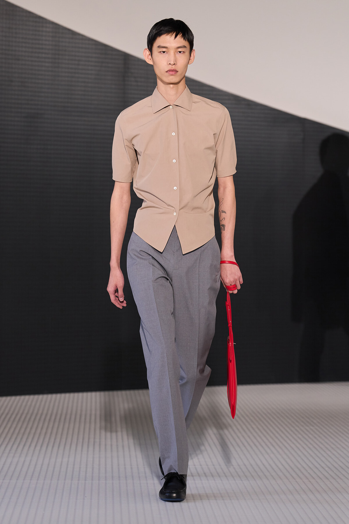 Male model on a runway wearing a beige short-sleeve button-up shirt and gray trousers, holding a red umbrella accessory.