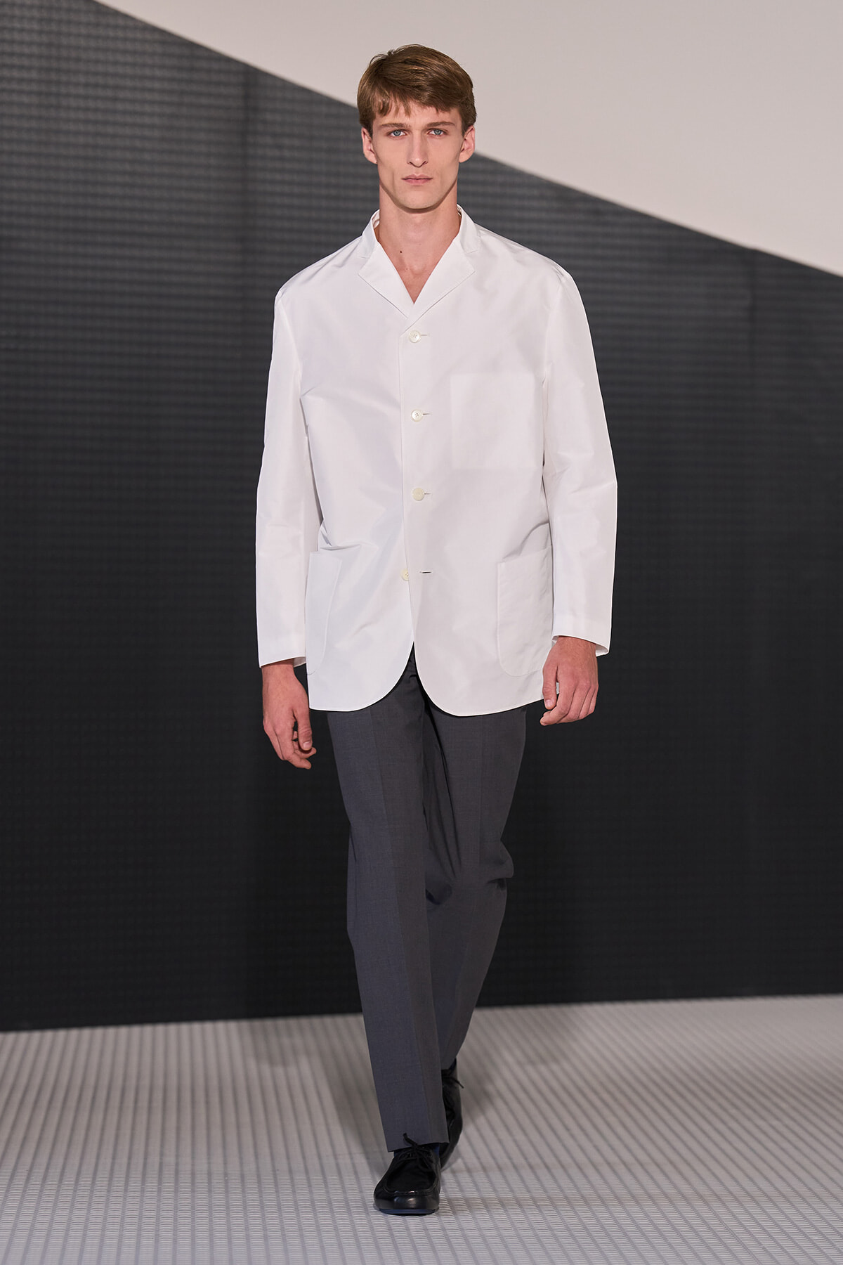 Male model walking on a runway in a white blazer with patch pockets and gray dress pants.