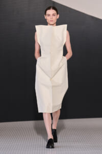 Runway model wearing a sculptural, off-white sleeveless dress with angular shoulders and a geometric silhouette.