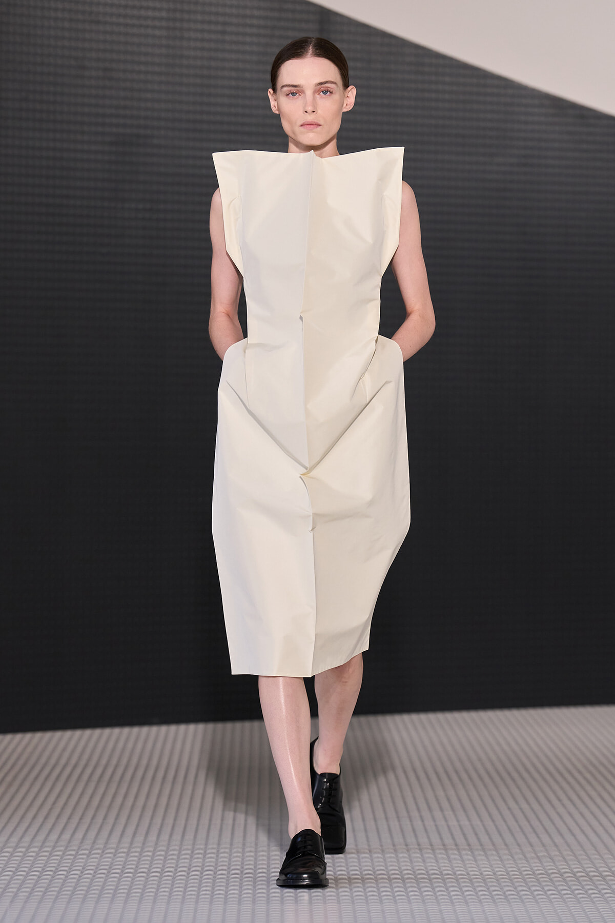 Runway model wearing a sculptural, off-white sleeveless dress with angular shoulders and a geometric silhouette.