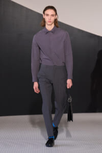 Male model on a fashion runway wearing a gray button-down shirt tucked into gray dress pants, black shoes, blue socks, and carrying a black bag with metal rings.
