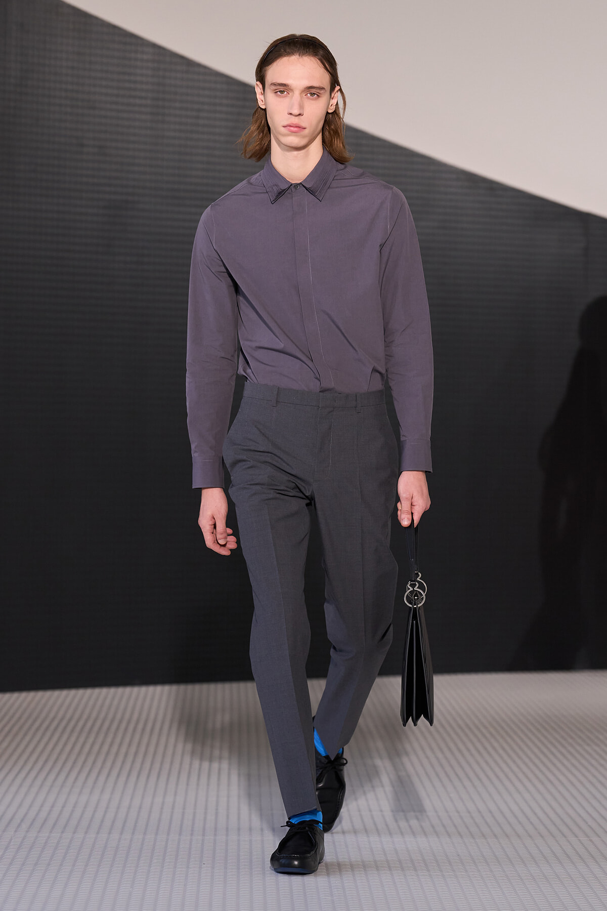 Male model on a fashion runway wearing a gray button-down shirt tucked into gray dress pants, black shoes, blue socks, and carrying a black bag with metal rings.