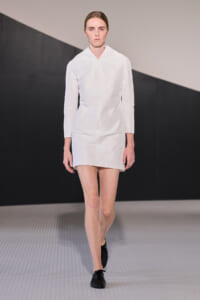 Runway model walking in a crisp white, minimalist knee-length dress with long sleeves and black shoes.