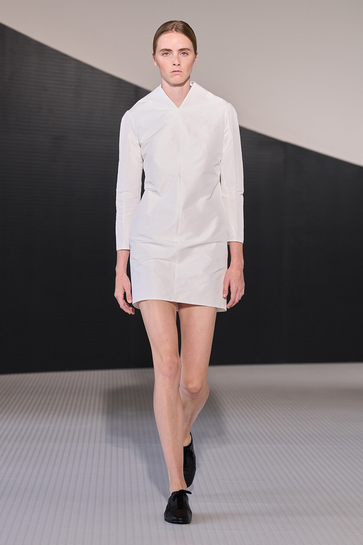 Runway model walking in a crisp white, minimalist knee-length dress with long sleeves and black shoes.