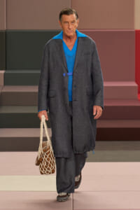 Male model in a dark gray long coat over a bright blue knit sweater, carrying a beige net tote on a fashion runway.