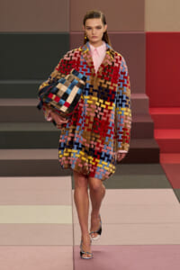 Model walking a runway in a bold multicolor patchwork coat, holding a matching woven tote bag under her arm.