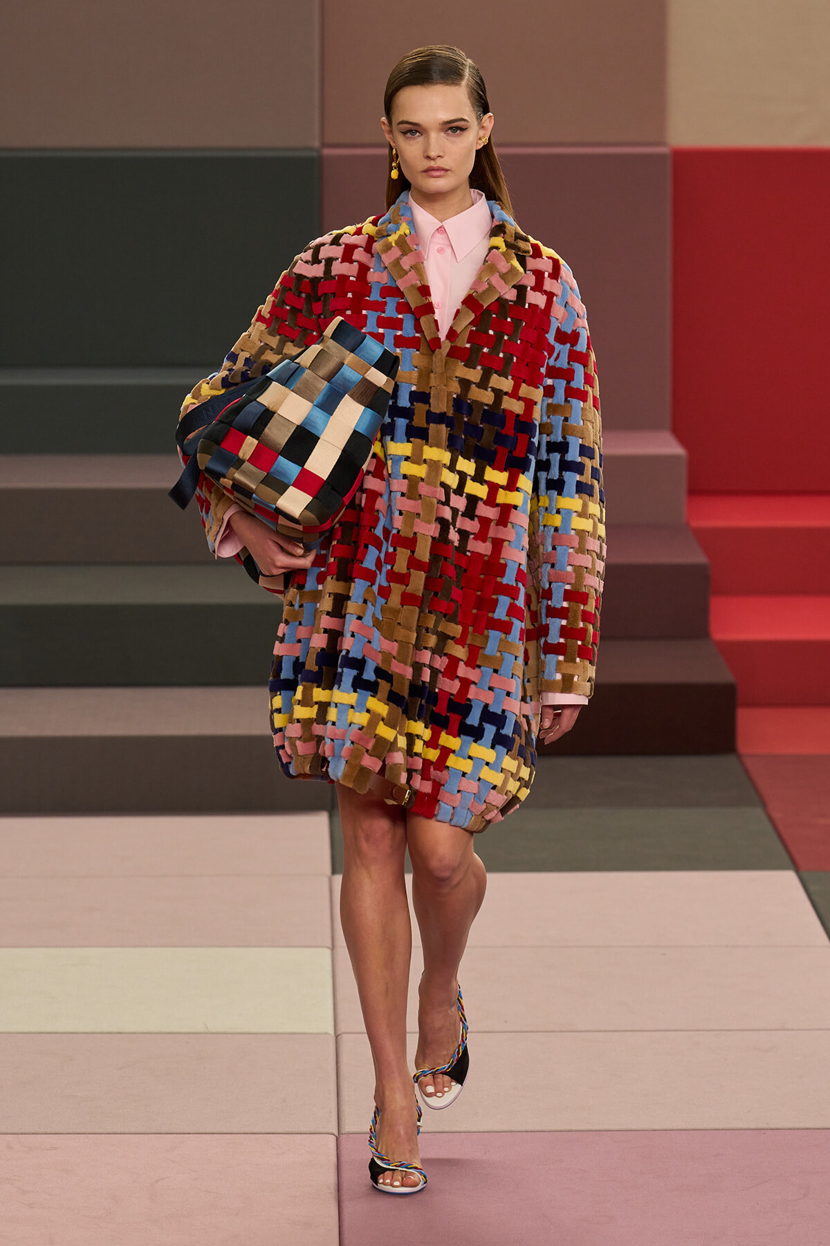 Model walking a runway in a bold multicolor patchwork coat, holding a matching woven tote bag under her arm.