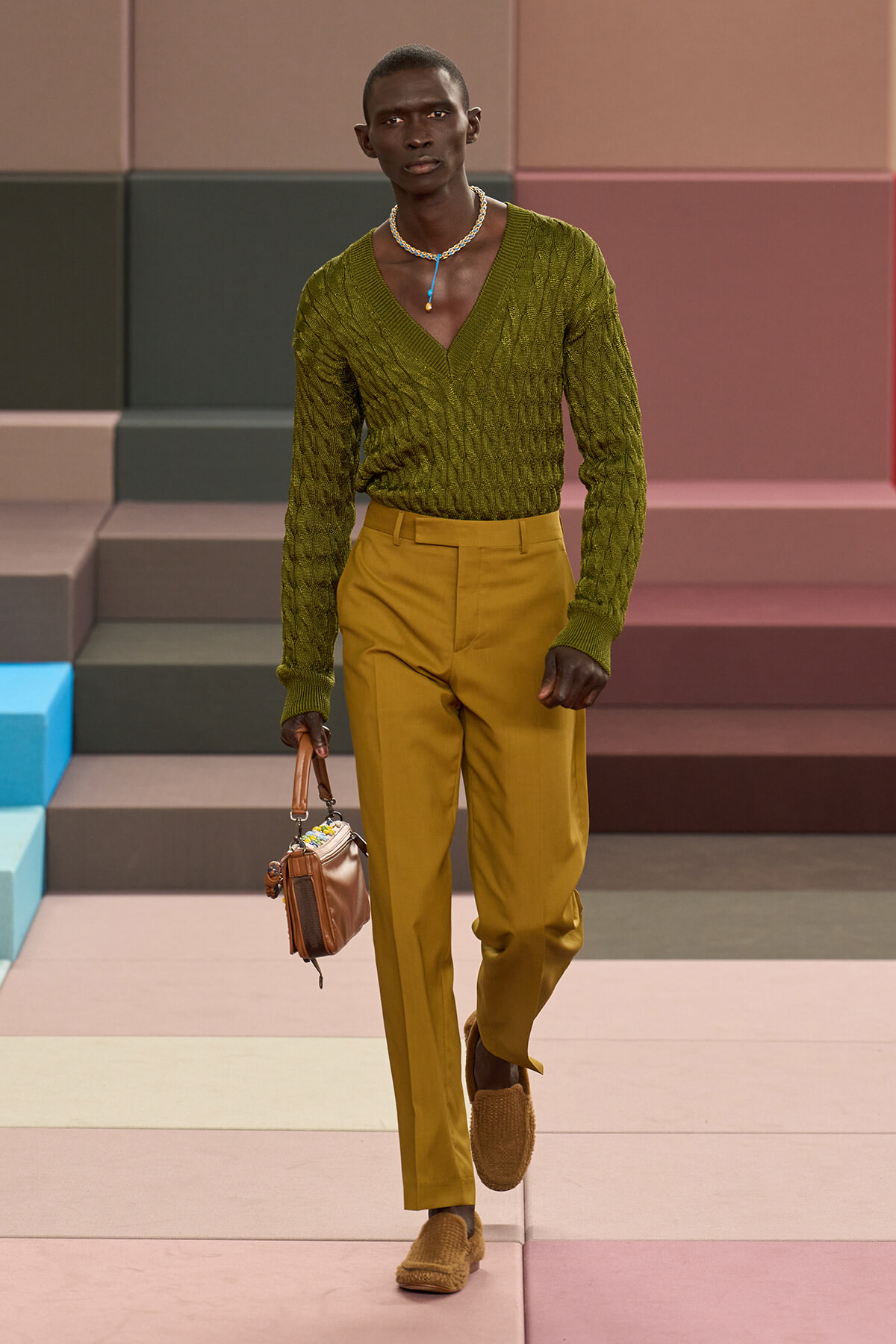 Male model walking runway in olive green V-neck sweater and mustard trousers, carrying a small tan handbag and wearing a beaded necklace and loafers.