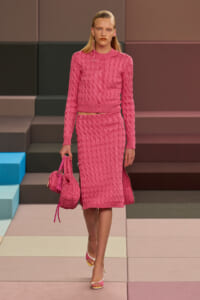 Model walks a runway in a pink cable-knit cardigan and matching midi skirt, carrying a pink quilted handbag.