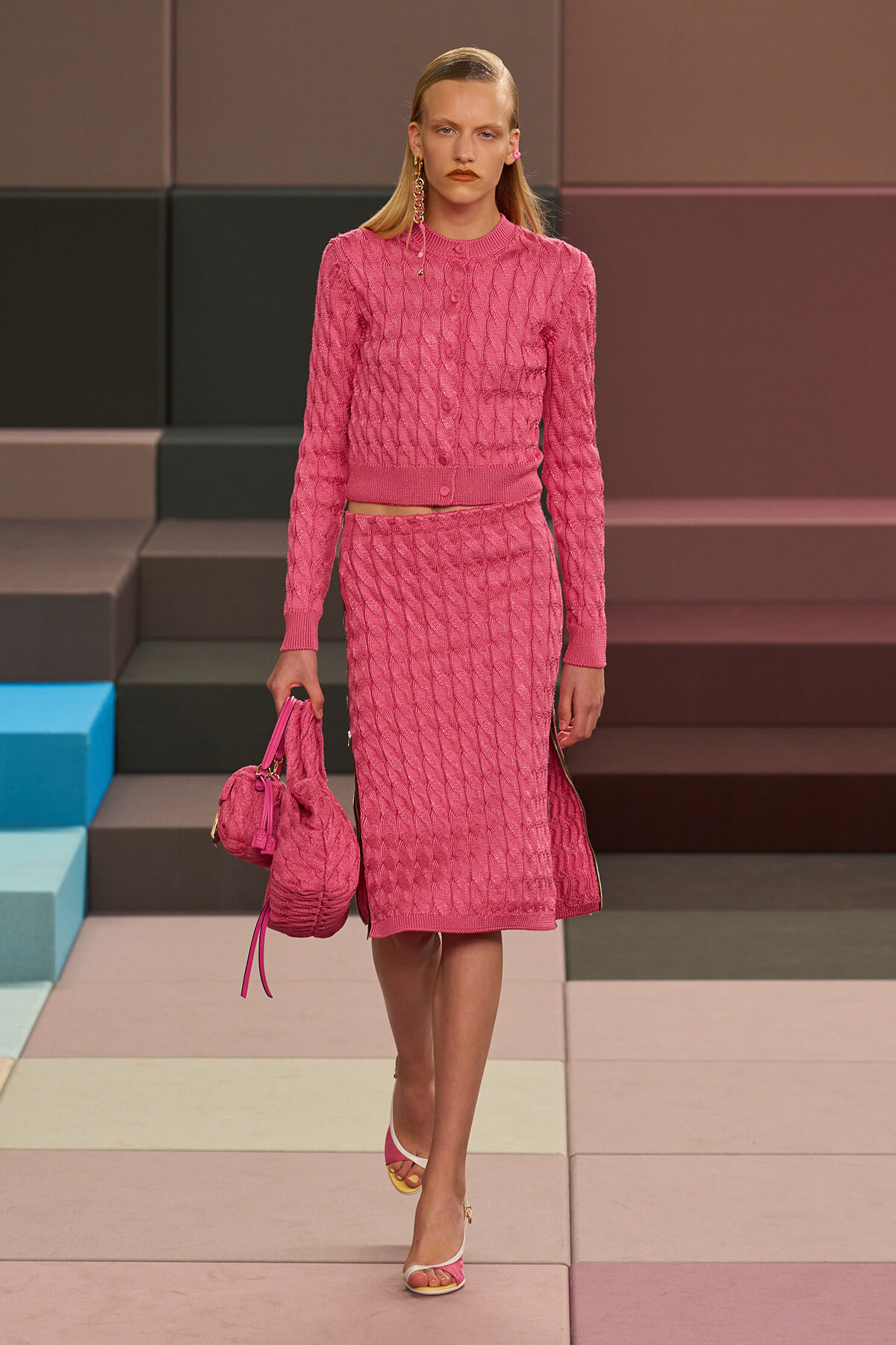 Model walks a runway in a pink cable-knit cardigan and matching midi skirt, carrying a pink quilted handbag.