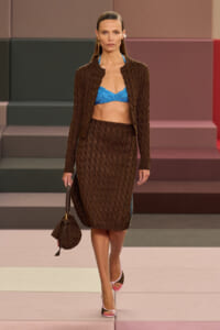 Model walking a runway in a textured brown knit suit with a blue bralette, carrying a brown knit handbag.