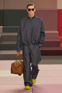 Model on a runway wearing a dark gray oversized zip-front jumpsuit with elastic waist, blue drawstrings, yellow sneakers, brown handbag, and sunglasses.