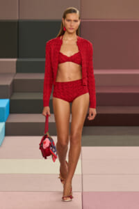 Model walking a runway in a matching red knit bikini and cardigan, carrying a red handbag with a scarf embellishment and wearing red sandals.