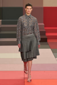 Model on a runway wearing a sheer gray lace blouse and a layered gray pleated skirt, mid-walk on a color-blocked set.