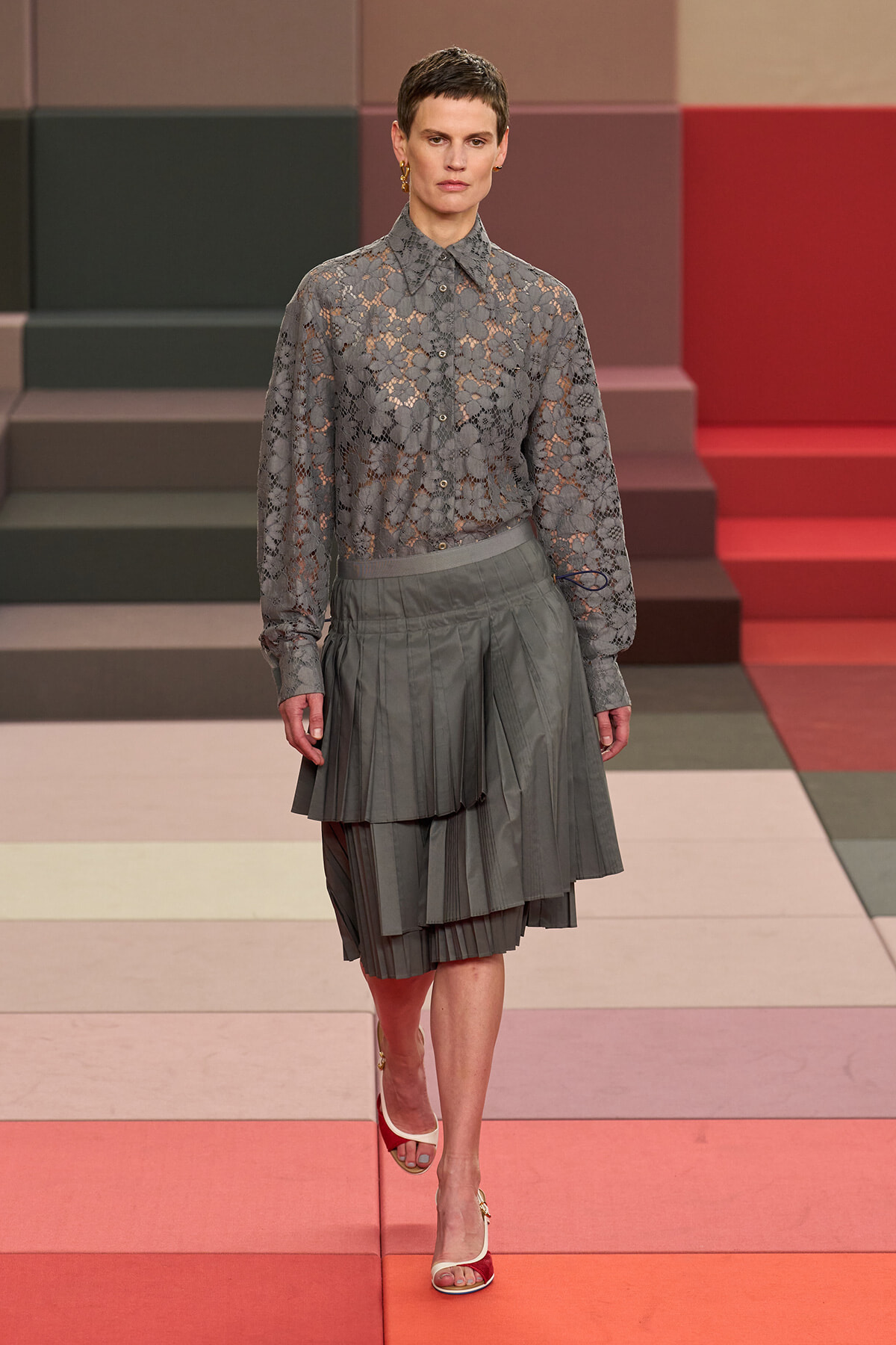 Model on a runway wearing a sheer gray lace blouse and a layered gray pleated skirt, mid-walk on a color-blocked set.