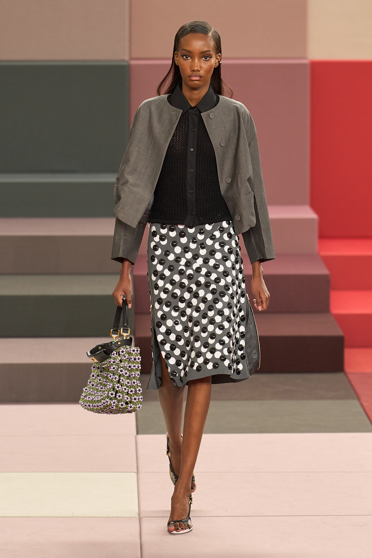 Model walking a fashion runway in a gray cropped jacket over a black knit blouse, paired with a gray midi skirt dotted with black circles; carries a beaded lavender-green handbag and wears strappy sandals.