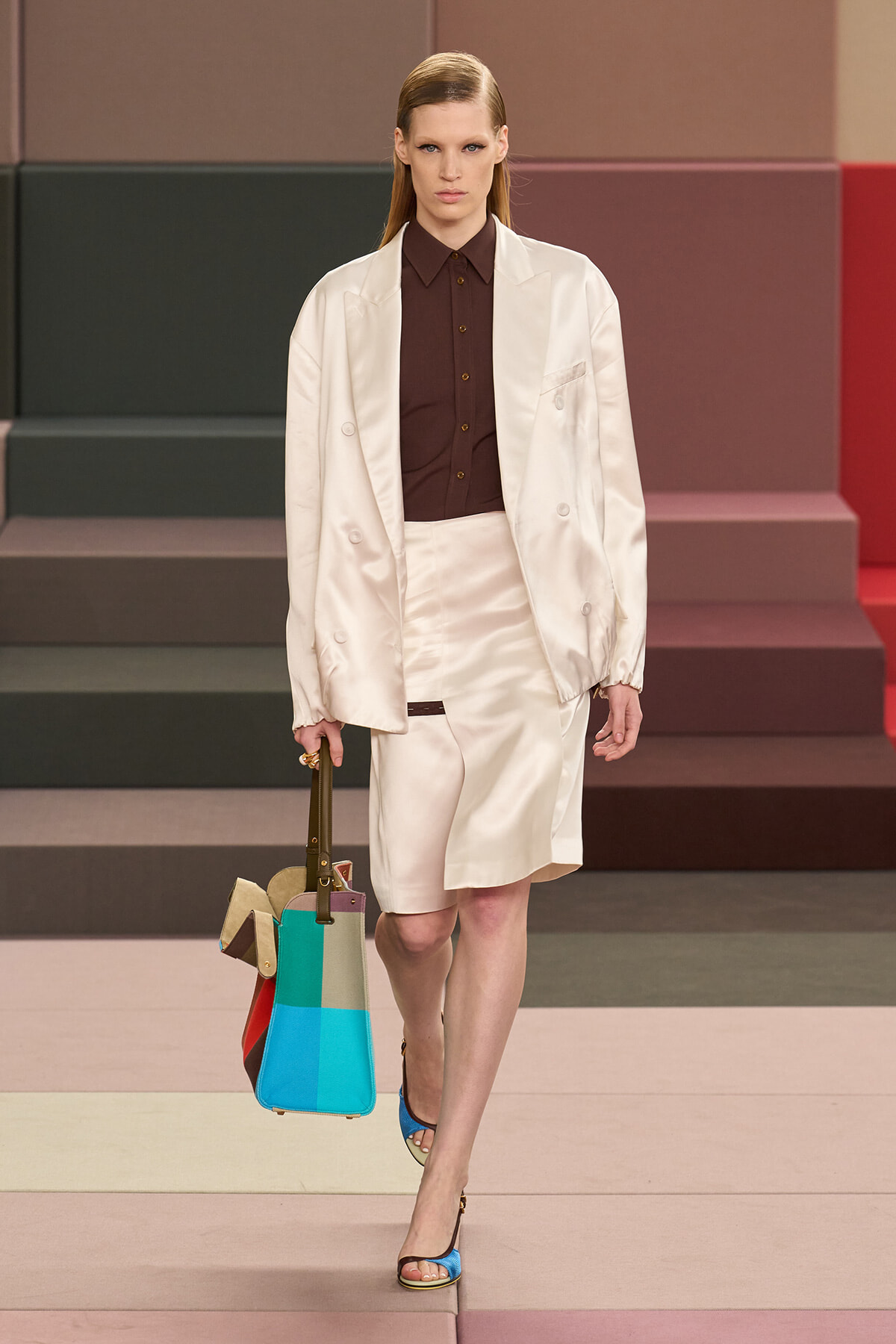 Model walking a runway in a cream suit with a brown shirt, carrying a color-block tote; blue sandals complete the look.