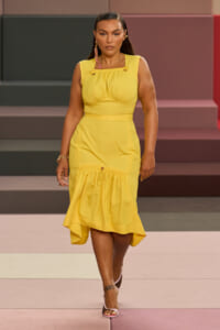 Model walking a runway in a bright yellow, sleeveless midi dress with ruched bodice and a ruffled hem, paired with pink-strap heels and gold jewelry.