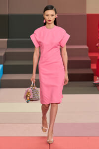 Model walks a pink knee-length dress with structured sleeves on a runway, carrying a beaded handbag with tassels.