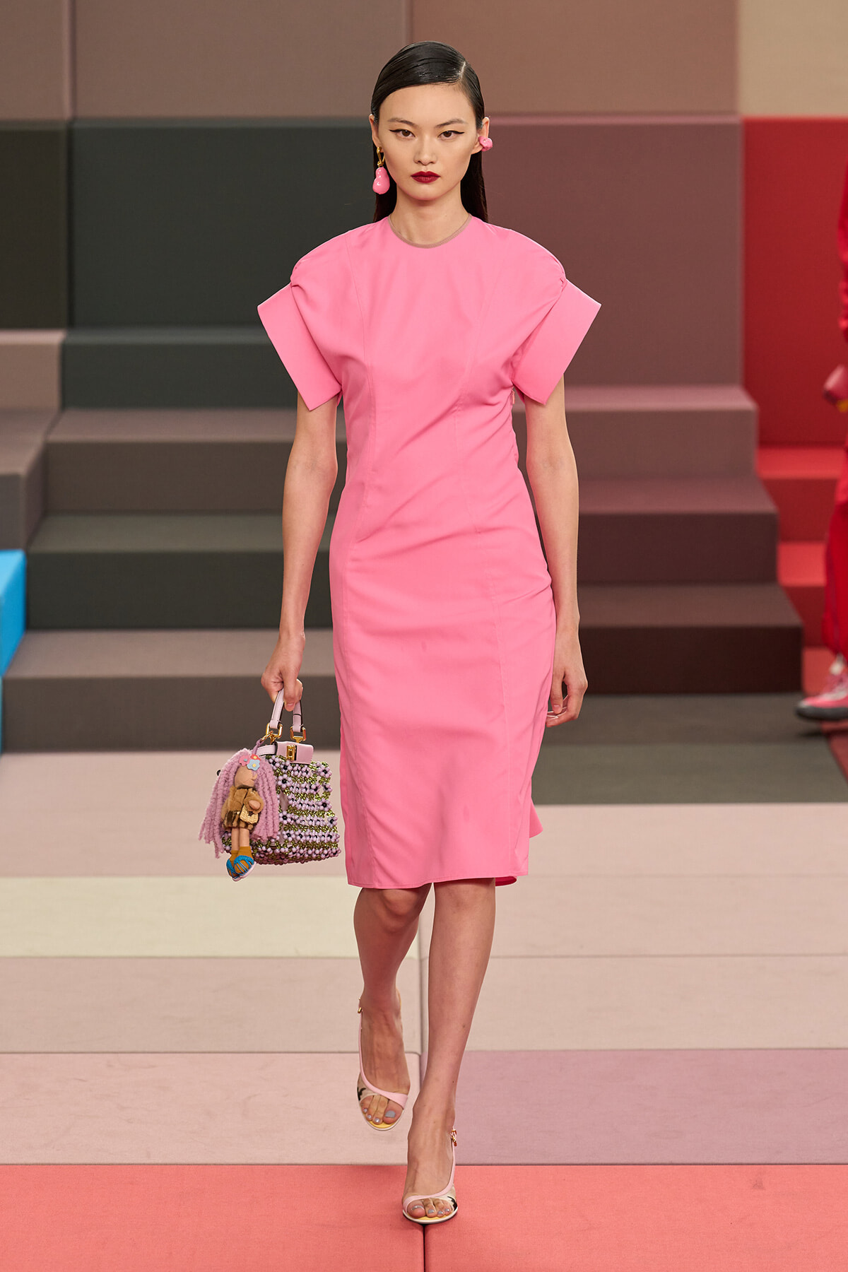Model walks a pink knee-length dress with structured sleeves on a runway, carrying a beaded handbag with tassels.