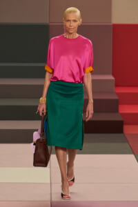 Model walking a runway in a bright pink satin top with orange cuffs and a green midi skirt, carrying a brown handbag and wearing pink sandals.