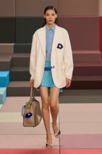 Model walking runway in oversized cream blazer over light blue shirt and turquoise pleated mini skirt, carrying a tan handbag with blue flower charm.