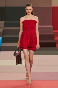Model walking a red strapless pleated mini dress on a runway, carrying a brown handbag and wearing gold sandals.