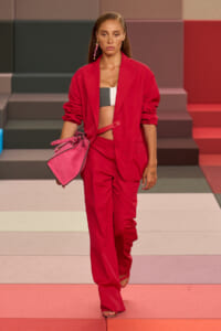 Model walking a fashion show in a red blazer and matching trousers, with a black-and-white crop top, carrying a pink handbag.