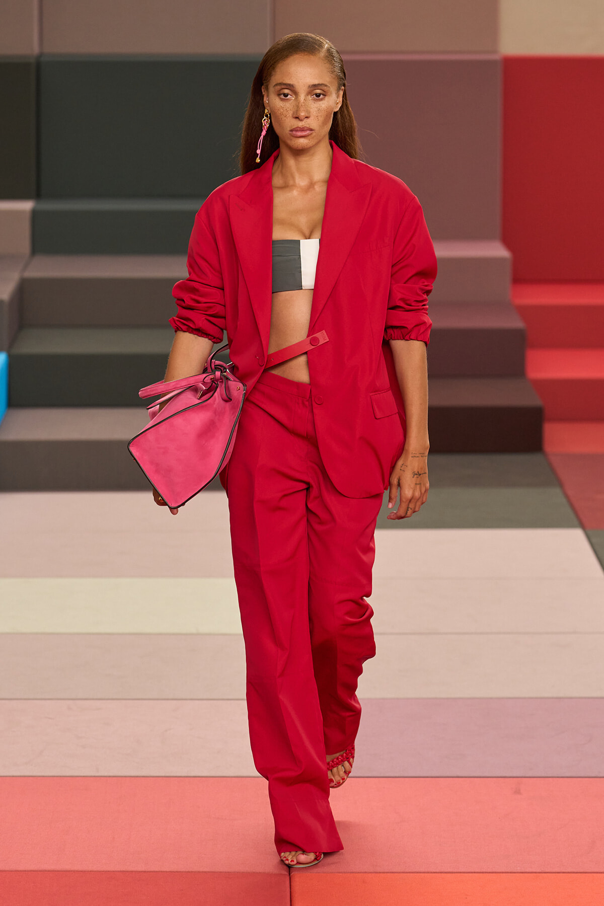 Model walking a fashion show in a red blazer and matching trousers, with a black-and-white crop top, carrying a pink handbag.