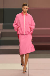 Model walking a runway in a pink zip-front windbreaker and matching pink midi skirt, wearing colorful braided heeled sandals.