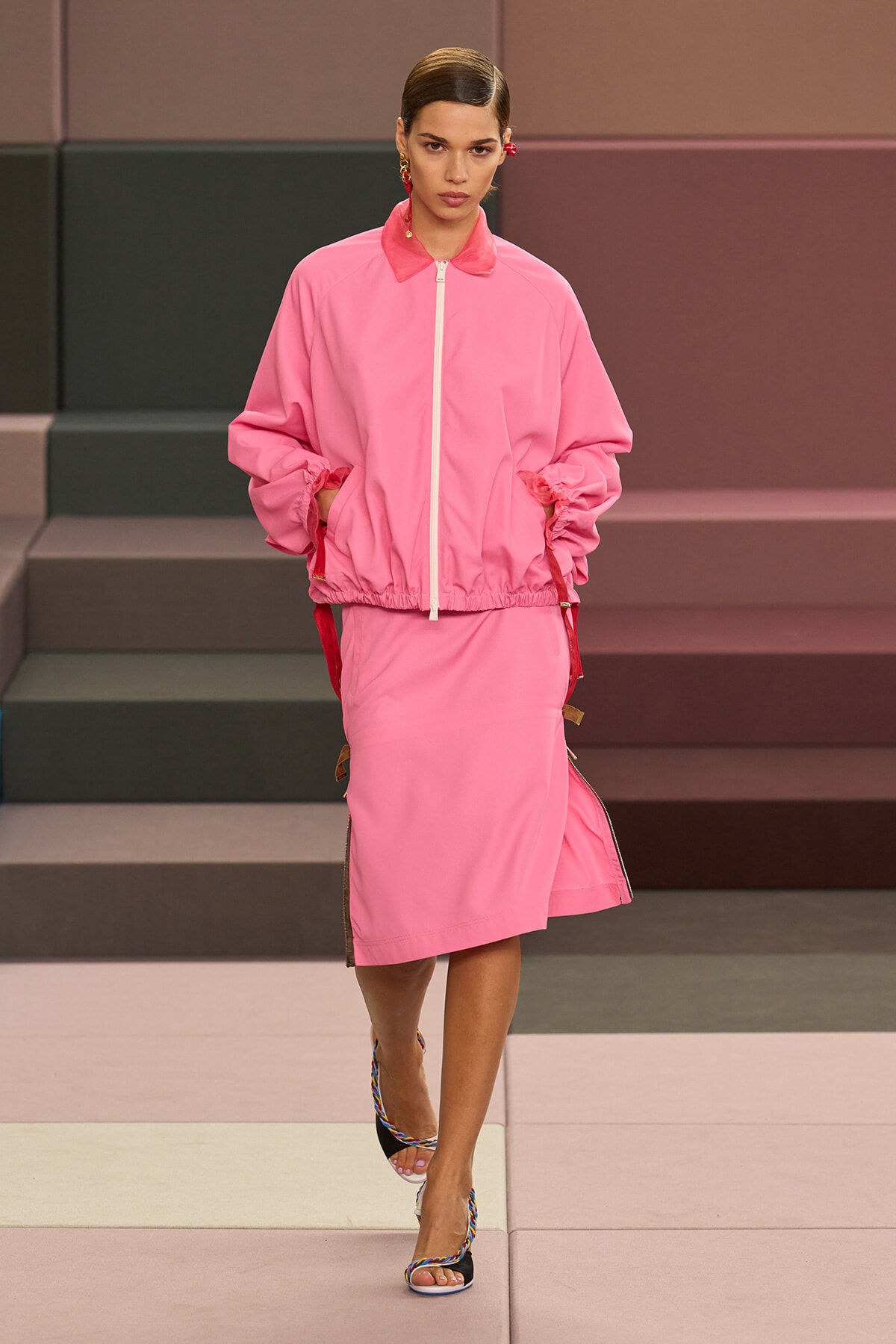Model walking a runway in a pink zip-front windbreaker and matching pink midi skirt, wearing colorful braided heeled sandals.