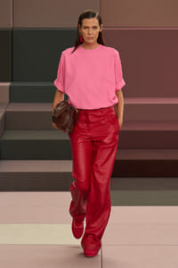 Model walking runway in bright pink blouse and red leather pants, holding a brown pouch-style bag