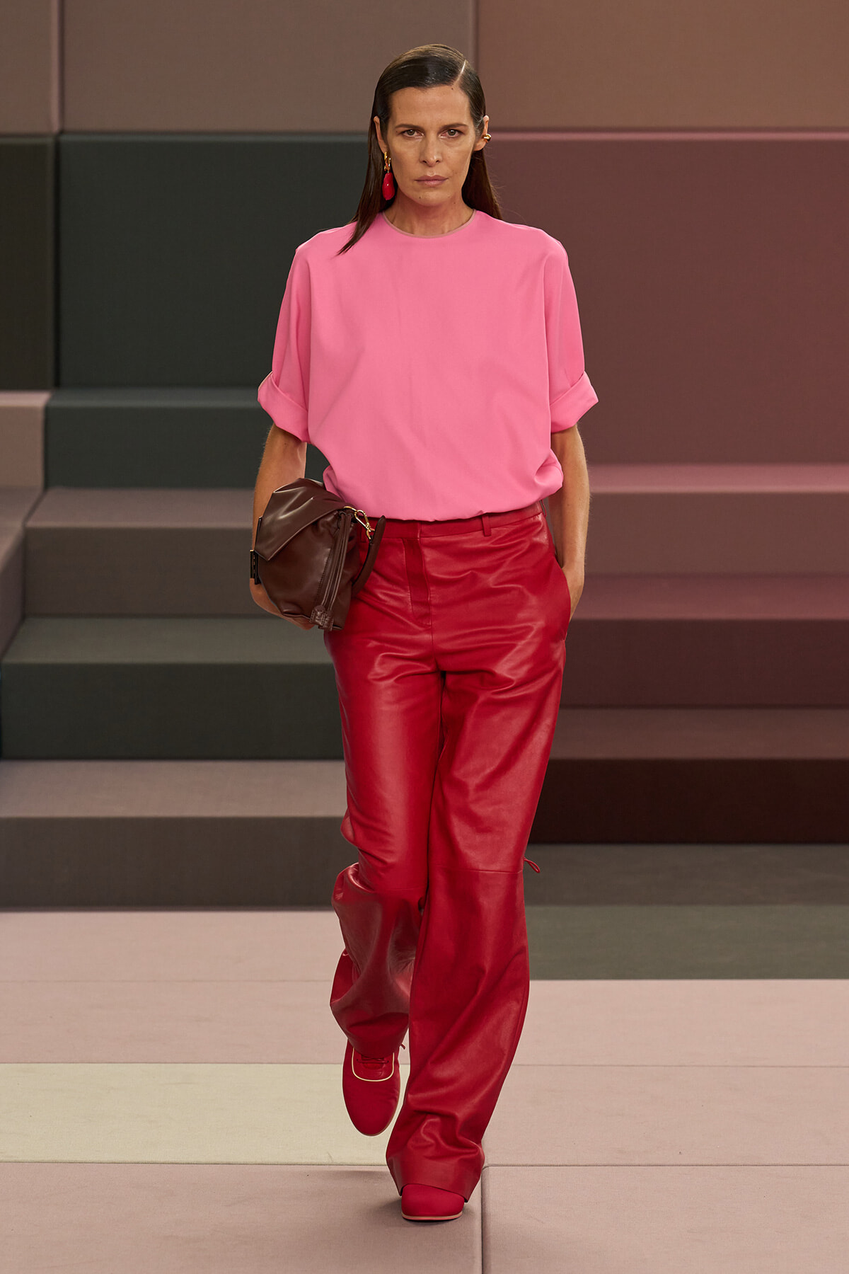 Model walking runway in bright pink blouse and red leather pants, holding a brown pouch-style bag