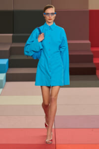 Model walking a fashion show in a bright blue button-down mini dress with oversized sleeves, blue sunglasses, and a matching blue handbag slung over the shoulder.