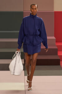 Model walking a runway in a navy belted tunic with a matching mini skirt, carrying a large white tote and wearing white strappy sandals.