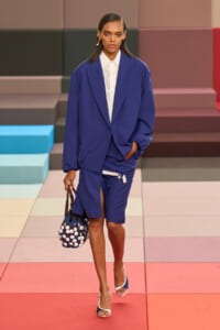 Model walks the runway in an oversized cobalt blue suit with a white buttoned blouse, carrying a navy polka-dot handbag and wearing white-and-blue heels on a pastel geometric floor backdrop.