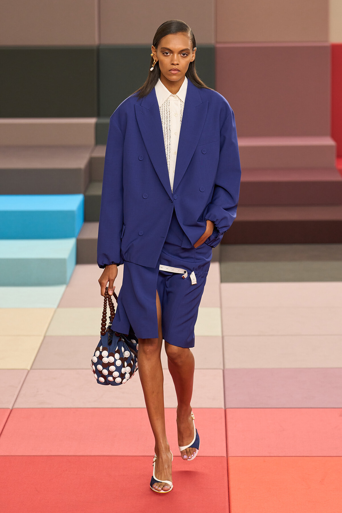 Model walks the runway in an oversized cobalt blue suit with a white buttoned blouse, carrying a navy polka-dot handbag and wearing white-and-blue heels on a pastel geometric floor backdrop.