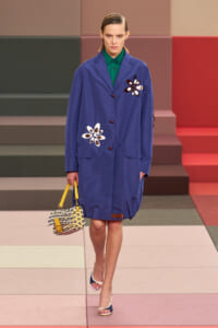 Model wearing a blue knee-length coat with white flower appliqués, green shirt, carrying a yellow-and-checked handbag on a patterned runway stage.