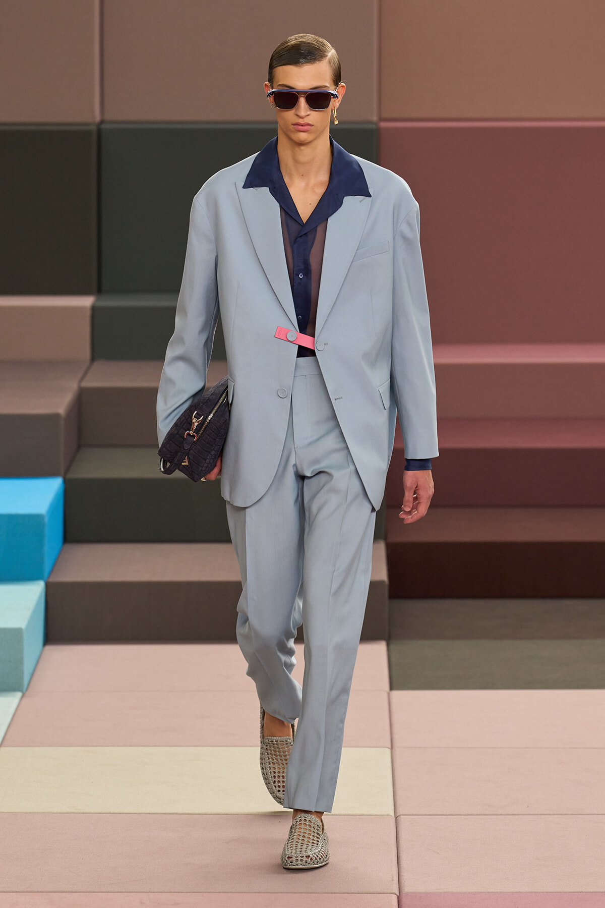 Runway model in a pale blue suit with navy shirt, pink belt, and sunglasses, carrying a black clutch on a pastel runway.