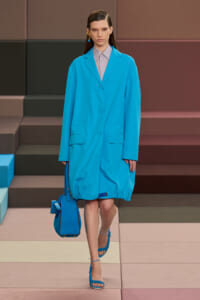 Model walking a runway in a bright turquoise oversized coat, lavender shirt, matching blue handbag and strappy blue sandals.