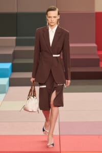 Model walking a runway in a brown suit with a white belt, white shirt, matching tailored blazer and split skirt; white bag with wooden bead handle hangs at her side.